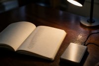 Geöffnetes Notizbuch mit handschriftlicher Zahlenreihe neben einem kleinen Metallgehäuse auf einem Tisch - Auf einem dunklen Holztisch liegt ein aufgeschlagenes Notizbuch mit einer handschriftlichen Reihe unleserlicher Wörter, daneben ein kleines silbernes Metallgehäuse mit Anschlusskabel und eine Leselampe. Symbolbild zur Aufbewahrung digitaler Zugangsschlüssel außerhalb von Handelsplattformen. Bild KI-generiert