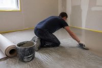 Handwerker spachtelt einen Untergrund vor der Bodenverlegung. Ein Handwerker zieht mit einer Glättkelle Spachtelmasse über einen Estrich, daneben stehen ein Eimer und aufgerollte Bodenbahnen. Symbolbild zum Markt für Verlegewerkstoffe und zur Sanierung im Bestand.