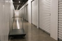 Gang mit verschlossenen Lagerabteilen in einer Selfstorage-Anlage. Ein langer, nüchtern beleuchteter Gang mit gleichförmigen verschlossenen Lagerabteilen und Rolltoren in einer Selfstorage-Anlage, im Vordergrund ein abgestellter Transportwagen. Symbolbild zum deutschen Markt für Lagerflächen zur Selbsteinlagerung.