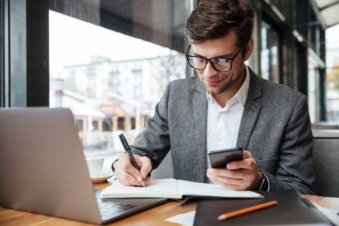 smiling-businessman-eyeglasses-sitting-by-table-cafe-with-laptop-computer-while-using-smartphone.jpg