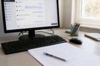 Bildschirm mit einem Chatfenster und einer daneben liegenden Liste offener Kundenfragen - Ein Monitor auf einem Schreibtisch zeigt ein geöffnetes Chatfenster einer Website, daneben liegt eine handschriftlich ergänzte Liste offener Kundenfragen und ein Kugelschreiber. Symbolbild zum maschinell unterstützten Kundendialog auf Unternehmensseiten.
