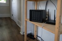 Kompakter Netzwerkspeicher mit vier Einschüben neben einem Router im Wohnungsflur. Ein kleines Speichergehäuse mit vier herausnehmbaren Festplatteneinschüben steht auf einem Holzregal, daneben ein Router mit angeschlossenem Netzwerkkabel und eine Steckdosenleiste. Symbolbild zur Datensicherung privater Haushalte im Heimnetz.