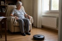 Ältere Frau in ihrer Wohnung neben einem Reinigungsroboter und einer Notrufuhr. Eine ältere Frau sitzt in ihrem Wohnzimmer in einem Sessel und trägt eine Uhr am Handgelenk, während ein flacher Reinigungsroboter über den Boden fährt. Symbolbild zu altersgerechten Assistenzsystemen im Haushalt.
