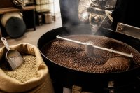 Geröstete Kaffeebohnen laufen aus der Kühltrommel einer kleinen Rösterei in einen Auffangbehälter. Die geöffnete Kühlschale einer kleinen Trommelrösterei, frisch geröstete Bohnen werden vom Rührarm verteilt, daneben ein Jutesack mit Rohkaffee und eine Schaufel, warmes Werkstattlicht. Symbolbild zum Rekordabsatz ganzer Bohnen und zur EU-Entwaldungsverordnung für Kaffee.