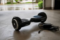 Hoverboard mit Ladegerät auf dem Boden einer Garage. Ein Hoverboard steht auf dem Betonboden einer Garage, daneben liegt ein Ladegerät mit aufgerollter Zuleitung. Symbolbild zum Umgang mit Akkus in Freizeitgeräten. Bild KI-generiert