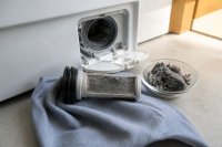 Nahaufnahme der unteren Wartungsklappe einer weißen Frontlader-Waschmaschine in einem Hauswirtschaftsraum, das Flusensieb ist herausgezogen und liegt auf einem Tuch, daneben eine kleine Schale mit gesammelten grauen Textilfasern, warmes Tageslicht von der Seite. Symbolbild zur Diskussion um Mikroplastikfilter in Waschmaschinen.