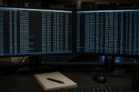 Zwei Bildschirme mit Transaktionslisten in einem abgedunkelten Büro - Zwei nebeneinanderstehende Monitore zeigen lange Listen aus Zeichenfolgen und Zeitstempeln, davor liegen ein Notizblock und ein Stift, der Raum ist abgedunkelt. Symbolbild zur Nachverfolgung von Transfers digitaler Vermögenswerte. Bild KI-generiert
