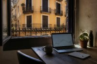 Ein schlichter Schreibtisch am Fenster einer Altbauwohnung mit Blick auf eine sonnige Straßenfassade, darauf ein aufgeklappter Laptop mit einem Liniendiagramm, ein Notizblock und eine Kaffeetasse. Symbolbild zum privaten Anlegen in Kryptowerte.