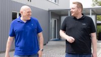 Zwei Männer in Poloshirts gehen lächelnd nebeneinander über das Außengelände eines Unternehmensgebäudes und unterhalten sich.