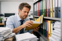 Mitarbeiter arbeitet mit papierbasierten Dokumenten und durchsucht Ordner in einem Archiv. Das Bild zeigt typische Herausforderungen manueller Dokumentenprozesse wie Zeitaufwand, fehlende Übersicht und ineffiziente Abläufe im Arbeitsalltag von Unternehmen.