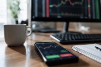 Smartphone mit geöffneter Handels-App und Kurskurve liegt neben Tastatur und Kaffeetasse auf einem Schreibtisch - Auf einem Schreibtisch liegt ein Smartphone mit einer geöffneten Handels-App, auf dem Bildschirm eine unscharfe Kurskurve und ein Kaufknopf, daneben eine Tastatur, eine Kaffeetasse und ein Notizblock; im Hintergrund ein Monitor mit Orderbuch. Symbolbild zum Verbot von Rückvergütungen für Wertpapieraufträge. Bild KI-generiert