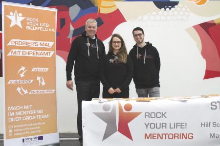 Vanessa Neumann (Mitte) und Tim Wagner (rechts) freuen sich über den Besuch von Holger Piening (links) an ihrem Stand in der Uni Bielefeld
