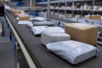 Kleine Versandkartons mit Zollaufklebern auf einem Sortierband in einer Paketverteilhalle - Mehrere kleine Versandkartons und gepolsterte Umschläge mit aufgeklebten Zollvermerken liegen auf einem Förderband in einer Paketverteilhalle, im Hintergrund unscharf Regale mit weiteren Sendungen. Symbolbild zum Pauschalzoll auf Kleinsendungen aus Drittstaaten. Bild KI-generiert