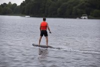 Fahrer auf einem elektrischen Surfboard mit Rettungsweste auf einem Badesee. Ein Fahrer steuert ein elektrisches Surfboard über einen ruhigen Badesee und trägt dabei eine Rettungsweste, am bewaldeten Ufer im Hintergrund liegen einzelne Boote. Symbolbild zur Sicherheit im motorisierten Wassersport auf Binnengewässern. Bild KI-generiert