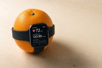 Symbolbild: Eine Smartwatch mit angezeigtem Blutdruckwert liegt um eine Orange — reale Smartwatch-Funktionen wie die Hypertonie-Benachrichtigung geben keinen mmHg-Wert aus und erreichen laut Herstellerangabe eine Sensitivität von 41,2 Prozent.