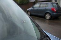 Steinschlag in einer Windschutzscheibe wird vor der Reparatur geprüft. Nahaufnahme einer Windschutzscheibe mit einem kleinen Steinschlag, dahinter unscharf ein älteres Fahrzeug in einer Hofeinfahrt. Symbolbild zum steigenden Durchschnittsalter der Fahrzeuge und zur wachsenden Bedeutung kleiner Reparaturen. Bild KI-generiert