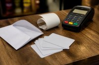 Auf einer hölzernen Ladentheke liegen mehrere unbedruckte Plastikkarten in Kartenformat, ein aufgeklappter Papierumschlag, ein Kassenbon und ein Kartenlesegerät. Symbolbild zum Markt für Gutscheinkarten und zum Einlöseverhalten im Handel.