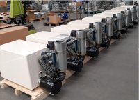 Das Bild zeigt eine Reihe von Schallschutzkabinen, die offenbar für den Versand oder die Auslieferung vorbereitet sind. Jede Kabine ist mit einem Seitenkanalverdichter ausgestattet, der über flexible Schläuche und ein Abgassystem verfügt. Die Kabinen sind auf Holzpaletten platziert, was auf eine geplante Logistik oder Lagerung hindeutet. Die Bauweise und die Anordnung der Komponenten zeigen, dass die Geräte für den Betrieb in einer schallgedämmten Umgebung konzipiert sind.