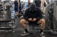 Trainierende in einer Fitnessanlage notieren ihre Einheit am Smartphone. Eine Person sitzt zwischen zwei Kraftgeraeten in einer Fitnessanlage und traegt die absolvierte Trainingseinheit in eine App auf dem Smartphone ein, im Hintergrund unscharf weitere Trainierende. Symbolbild zur wachsenden Zahl der Mitglieder in Fitness- und Gesundheitsanlagen. Bild KI-generiert