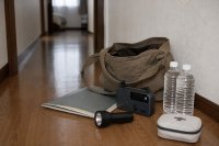 Gepackte Tasche mit Dokumentenmappe, Taschenlampe und Trinkwasser im Flur einer Wohnung. Eine offene Umhängetasche steht auf dem Boden eines Wohnungsflurs, daneben eine Dokumentenmappe, eine Taschenlampe, ein Kurbelradio, Wasserflaschen und ein kleiner Verbandkasten. Symbolbild zum griffbereiten Notgepäck in der privaten Krisenvorsorge.