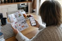 Person vergleicht am Küchentisch einen gedruckten Prospekt mit dem Smartphone