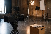 Eine digitale Filmkamera mit Cine-Objektiv auf einem Stativ vor einem leeren Set, im Hintergrund Scheinwerfer auf Stativen und eine Filmklappe auf einem Apfelkasten, warmes Arbeitslicht. Symbolbild zur Produktion europäischer Filme und Serien in Deutschland.