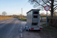 Am Rand einer zweispurigen Landstraße steht ein grauer Anhänger mit einer mobilen Messanlage, dahinter ein Verkehrsschild mit Tempolimit und die Einfahrt zu einem Schulgelände, die Fahrbahn ist leer. Symbolbild zur Verkehrsüberwachung und zu Kontrollwochen der Polizei.