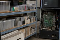Ein Depotregal mit sortierten Spielmodulen, Disketten und Handbüchern steht neben einem geöffneten Arcade-Automatengehäuse mit sichtbarer Platine. Symbolbild zur Archivierung von Computerspielen und Automatentechnik.
