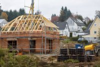 Rohbau eines Einfamilienhauses mit Gerüst und Dachstuhl in einem Neubaugebiet am Ortsrand. Ein frisch gemauerter Rohbau mit Gerüst und noch offenem Dachstuhl steht in einem Neubaugebiet, daneben ein Bauzaun, ein Kran und ein Stapel Dachziegel auf Paletten; im Hintergrund bestehende Wohnhäuser mit Gärten. Symbolbild zu den Baugenehmigungen für Wohnungen im Jahr 2026. Bild KI-generiert