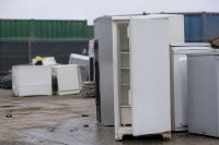 Ausgedientes Kühlgerät auf einem Sammelplatz für Elektroaltgeräte. Ein älteres weißes Kühlgerät steht mit offener Tür neben weiteren ausgedienten Haushaltsgeräten auf dem Sammelplatz eines Wertstoffhofs, im Hintergrund unscharf Container und Zaun. Symbolbild zur Verwertung von Elektroaltgeräten in Deutschland.