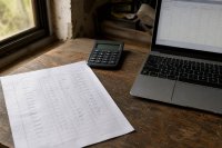 Handschriftlicher Stundenzettel neben Taschenrechner und Laptop auf einem Werkstatttisch