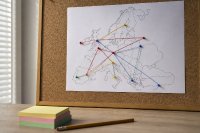 Europakarte auf einer Pinnwand mit farbigen Nadeln und verbindenden Fäden zwischen Standorten - Eine schlichte Europakarte hängt an einer Korkpinnwand, mehrere farbige Stecknadeln markieren verschiedene Standorte und sind durch gespannte Fäden miteinander verbunden, daneben liegen ein Notizzettelstapel und ein Stift auf einem Holztisch. Symbolbild zur grenzüberschreitenden Besetzung von Engpassberufen. Bild KI-generiert.