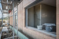 Ein Mehrfamilienhaus im Rohbau mit Gerüst an der Fassade, durch eine offene Fensteröffnung im Erdgeschoss ist eine frisch verputzte, noch feuchte Innenwand mit Putzprofilen zu sehen, davor Eimer und ein Glättbrett. Symbolbild zum Übergang vom Rohbau zum Ausbau.