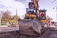 Ein Bagger von Rath arbeitet auf einer Straßenbaustelle in einem Wohngebiet. Der Fahrer in rotem Oberteil bedient die Maschine, während die Schaufel den Asphalt aufreißt. Im Hintergrund sind Absperrungen, herbstliche Bäume, ein Gartenzaun und ein Mobilfunkmast zu sehen. Die Szene vermittelt aktive Infrastrukturarbeit, etwa im Bereich Tiefbau oder Leitungserneuerung. Ein Bagger von Rath arbeitet auf einer Straßenbaustelle in einem Wohngebiet. Der Fahrer in rotem Oberteil bedient die Maschine, während die Schaufel den Asphalt aufreißt. Im Hintergrund sind Absperrungen, herbstliche Bäume, ein Gartenzaun und ein Mobilfunkmast zu sehen. Die Szene vermittelt aktive Infrastrukturarbeit, etwa im Bereich Tiefbau oder Leitungserneuerung.