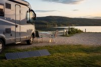 Ein Wohnmobil steht auf einem einfachen Stellplatz am Ufer eines Sees, davor ein aufgeklapptes tragbares Solarmodul auf dem Rasen, ein Campingtisch mit zwei Stühlen und ein leerer Stellplatz daneben im Abendlicht. Symbolbild zu Campingübernachtungen und zur Stromversorgung unterwegs im Sommer 2026. Bild KI-generiert.