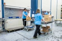 Mitarbeitende von BeeWaTec bei der Kommissionierung im Lager: Zwei Personen entnehmen Waren aus einem Hänel-Lagersystem und stellen die benötigten Artikel für Kundenaufträge zusammen. Im Vordergrund transportiert eine weitere Person einen Kommissionierwagen mit bereits verpackten Kartons durch den Lagerbereich. Die Bewegungsunschärfe verdeutlicht die Dynamik und Effizienz moderner Logistikprozesse.