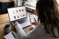 Person vergleicht am Küchentisch Produktangebote auf Laptop und Smartphone. Eine Person sitzt am Küchentisch und vergleicht Produktangebote gleichzeitig auf einem Laptop und einem Smartphone, daneben liegen eine Kaffeetasse und ein Notizzettel. Symbolbild zur wachsenden Bedeutung von Onlinemarktplätzen im deutschen Einzelhandel. Bild KI-generiert