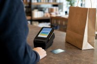 Kundin an der Kasse eines Ladengeschäfts wählt eine Ratenzahlung am Terminal. Eine Kundin steht an einem Ladentisch vor einem Kartenterminal, auf dem Display ist eine Auswahl von Zahlungsoptionen angedeutet, daneben liegen eine Einkaufstüte und eine Debitkarte. Symbolbild zum neuen Verbraucherkreditrecht. Bild KI-generiert