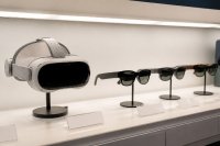Verkaufsregal in einem Elektronikgeschäft mit einem VR-Headset und mehreren brillenförmigen Smart Glasses auf Präsentationsständern - Ein beleuchtetes Verkaufsregal in einem Elektronikfachgeschäft: links ein kompaktes VR-Headset mit Kopfband auf einem Ständer, rechts drei brillenförmige Smart Glasses mit dunklen Gläsern auf kleinen Präsentationsständern, davor leere Preisschildhalter. Symbolbild zur Verschiebung des XR-Marktes von Headsets zu Smart Glasses im Jahr 2026. Bild KI-generiert.