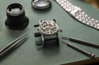 Eine Uhrmacher-Werkbank aus der Nähe: ein geöffnetes mechanisches Uhrwerk in einer Halterung, daneben Uhrmacherlupe, Pinzetten, feine Schraubendreher und ein Stahlarmband auf grünem Arbeitsleder, weiches Tageslicht von der Seite. Symbolbild zur Monatsstatistik der Schweizer Uhrenexporte im Oktober 2026.