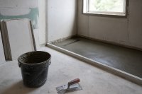 Rohbau einer bodengleichen Dusche mit Gefälleestrich, Ablaufrinne und gestapelten Fliesenkartons. Ein Badezimmer im Umbau: Der Boden einer bodengleichen Dusche ist mit Gefälleestrich und einer eingebauten Ablaufrinne vorbereitet, an der Wand stehen gestapelte Fliesenkartons, ein Zahnspachtel und ein Eimer mit Fliesenkleber, durch das Fenster fällt Tageslicht. Symbolbild zum Zuschuss der Pflegekasse für das barrierefreie Bad. Bild KI-generiert