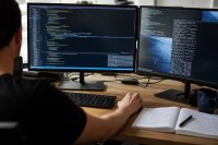 Entwicklerin prüft den Programmcode eines Finanzprotokolls an zwei Monitoren. Eine Entwicklerin sitzt an einem Schreibtisch vor zwei Monitoren und prüft Quellcode und Protokollausgaben eines Finanzprogramms, daneben liegen handschriftliche Notizen. Symbolbild zur Sicherheitsprüfung dezentraler Finanzanwendungen. Bild KI-generiert