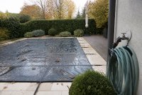 Abgedeckter Gartenpool im Herbst mit aufgerolltem Gartenschlauch und Laub auf der Plane. Ein rechteckiger Gartenpool ist mit einer dunklen Plane abgedeckt, auf der einzelne Herbstblätter liegen, daneben ein aufgerollter Gartenschlauch an einem Außenwasserhahn und eine Terrasse im Hintergrund. Symbolbild zur Poolbefüllung, zum Wasserverbrauch von Gartenpools und zur Winterpause. Bild KI-generiert