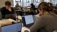 Studierende arbeiten konzentriert an Laptops und Büchern in einer Bibliothek oder einem Lesesaal. Es herrscht eine stille, fokussierte Atmosphäre.