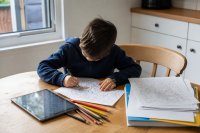 Ein Grundschulkind sitzt am Küchentisch und malt ein ausgedrucktes Ausmalbild mit Buntstiften aus, daneben liegen Papierbögen und ein abgeschaltetes Tablet. Symbolbild zum Spielverhalten von Kindern zwischen Bildschirm und Papier.