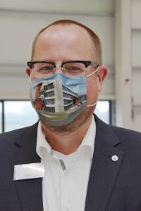 So sieht Vorfreude aus: AVENTUS-Geschäftsführer Kai Lammers trägt einen Mund-Nase-Schutz, bedruckt mit einer Visualisierung des AVENTUS-Neubaus.
