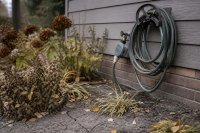 Aufgerollter Gartenschlauch mit Bewässerungscomputer am Wasserhahn vor einem herbstlichen Beet mit rissigem Boden - Ein aufgerollter Gartenschlauch hängt an einer Hauswand, am Außenwasserhahn sitzt ein kleiner Bewässerungscomputer, davor ein herbstliches Beet mit trockenem, rissigem Boden und ersten gefallenen Blättern. Symbolbild zur Gartenbewässerung am Ende der Saison 2026 nach dem Dürresommer. Bild KI-generiert.