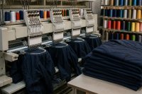 In einer Werkstatt für Textilveredelung läuft eine industrielle Mehrkopf-Stickmaschine, in den Rahmen sind dunkelblaue Arbeitsjacken eingespannt, daneben stehen Garnkonen in mehreren Farben auf einem Ständer und gestapelte fertige Jacken auf einem Tisch. Symbolbild zur Textilkonjunktur und zur Auftragslage in der Textil- und Bekleidungsindustrie im Herbst 2026.