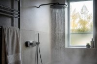 Laufende Dusche mit digitaler Temperaturanzeige an der Armatur in einem hellen Badezimmer. In einem hellen Badezimmer läuft Wasser aus einer Regendusche, an der Wandarmatur leuchtet eine kleine digitale Anzeige mit unleserlichen Ziffern, daneben ein Handtuch auf einem Heizkörper. Symbolbild zur Warmwasserbereitung und zum Wasser- und Energieverbrauch im Bad. Bild KI-generiert
