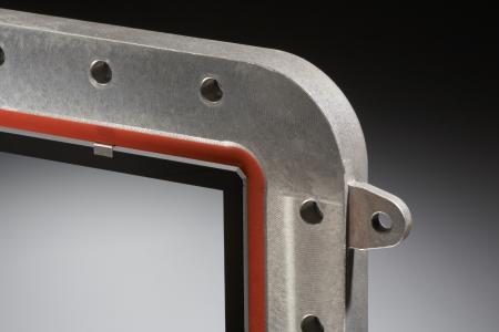 Einbaufertige Sicht- und Schutzscheibe. Die transparente und beschichtete Glaskeramik ist in einen Rahmen aus Aluminium-Druckguss eingeklebt. Die Glaskante ist umlaufend facettiert und ohne scharfe Kanten, um in der Einbausituation Platz für die Dichtung (rot) zu schaffen. Die elektrisch leitende Beschichtung ist mittels Kontaktfedern angebunden (im Bild oben links).
© Irlbacher